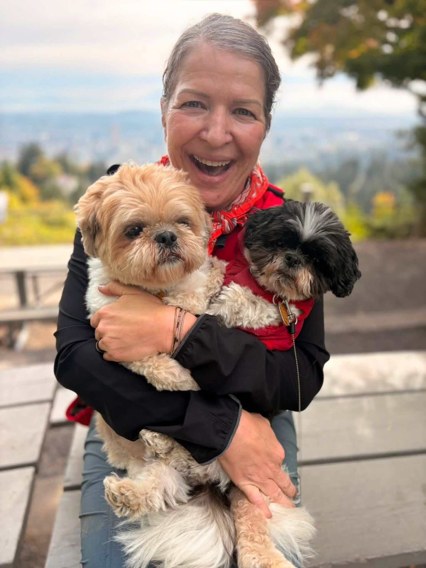 Crystal Mattox laughing outdoors, hugging her two small fluffy dogs, with trees and a city view behind her